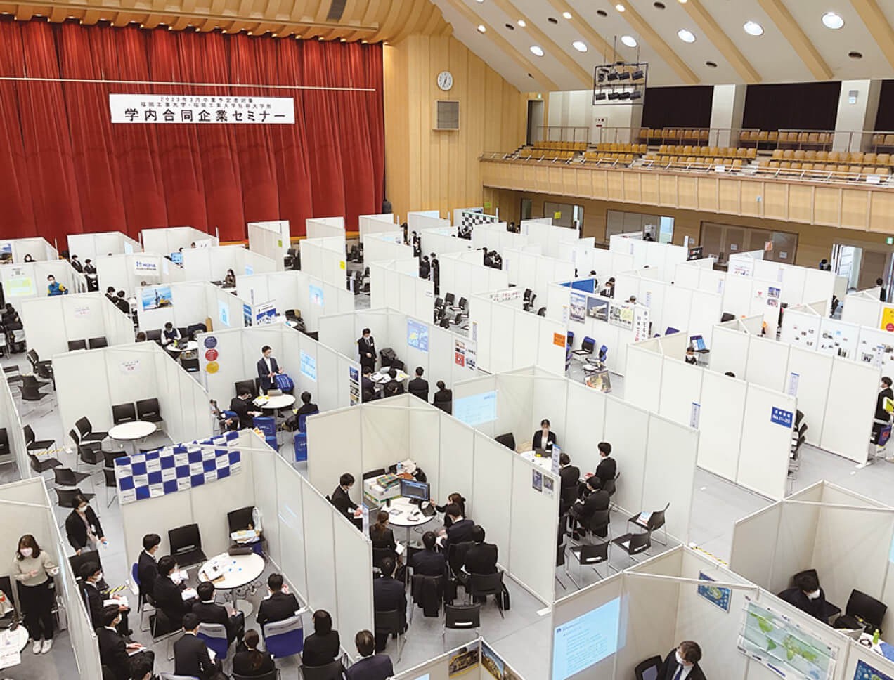 写真：福岡工業大学内で行われている、大学独自の合同企業セミナーの様子。スーツを来た学生が各企業ブースで説明を受けている。